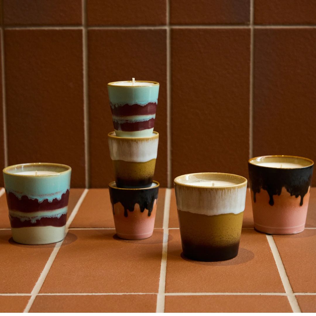 Flere HK Living duftlys i 70s Ceramics-krus i ulike farger og glasurer, vist samlet på et terrakottafarget flisgulv.