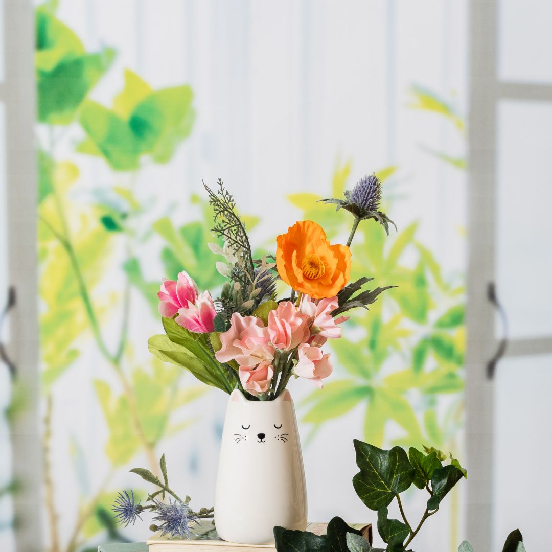hvit kattevase med blomster på bord ved vindu